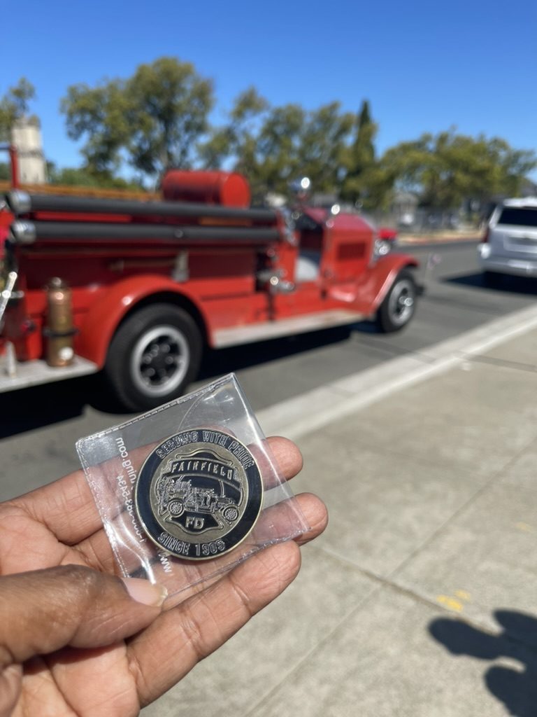 Commemorative coin held up in front of a vintage red fire engine at the Fairfield Fourth of July Parade